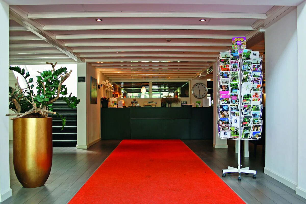 Vestíbulo de hotel con alfombra roja, mostrador de recepción, una planta y una exposición de fotografías.