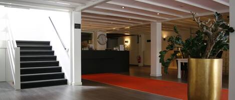 A hotel lobby with a staircase, a reception desk, a large potted plant, and a red carpet.