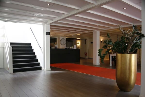 A hotel lobby with a staircase, a reception desk, a large potted plant, and a red carpet.