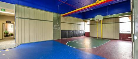 An indoor basketball court with a blue floor, a basketball hoop, and a wall with a warning sign.