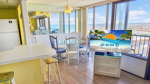 Una cocina con un ventanal amplio, una zona de comedor con sillas, un refrigerador y un televisor mostrando una escena de playa.