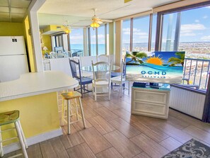 Eine Küche mit einem großen Fenster, einem Essbereich mit Stühlen, einem Kühlschrank und einem Fernseher, der eine Strandansicht zeigt.