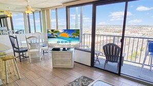 Un balcón con vista a la ciudad, un televisor sobre un mueble, sillas blancas y una alfombra estampada.