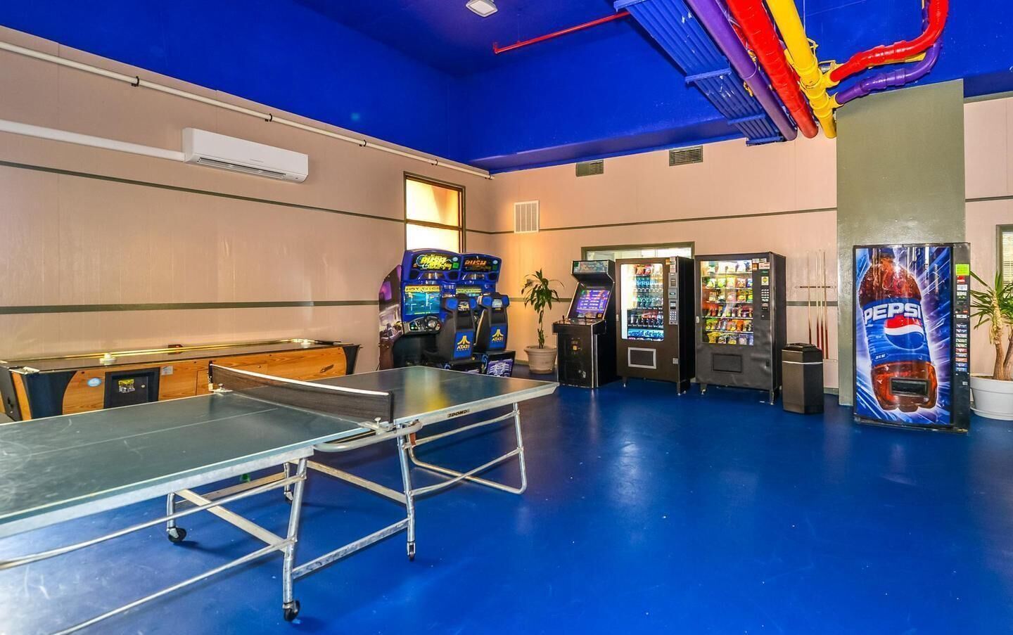 A ping pong table, vending machines, and arcade games in a room with blue flooring and a ceiling with colorful pipes.