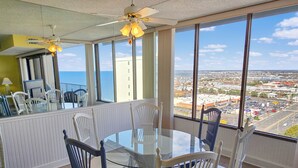 Un comedor con una mesa y sillas de vidrio, ventanas amplias con vistas a la ciudad y un ventilador de techo.