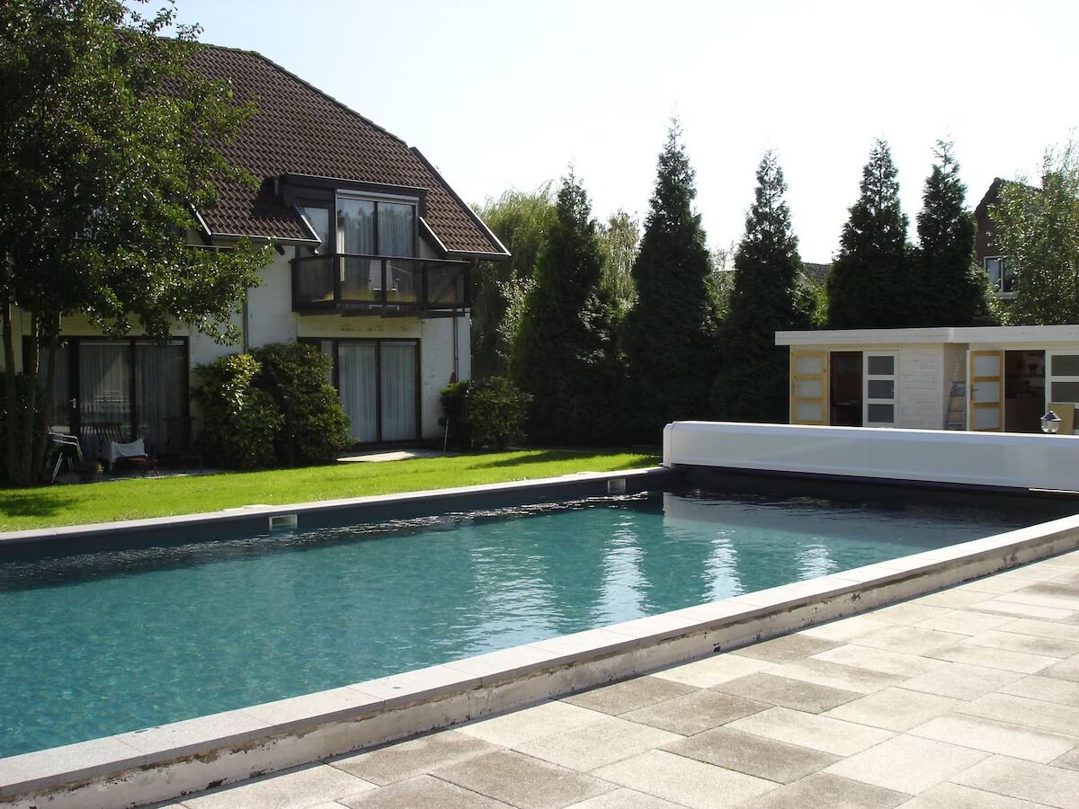 Een zwembad met betegeld zonnedek, een huis met schuin dak en een gazon met bomen.