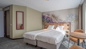A hotel room with a bed, a small table, a chair, a mirror, and a floral mural on the wall.