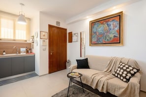 Uma sala de estar moderna com sofá, uma mesa pequena, um quadro e uma área de cozinha com armários e pia.