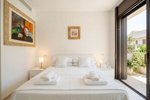 Quarto de hotel com uma cama grande, duas mesas de cabeceira, um quadro na parede e vista através de uma porta de vidro.