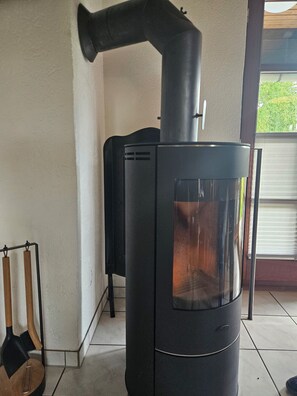 Un estufa de leña negra con puerta de vidrio, una chimenea de metal y un soporte metálico con un gancho.