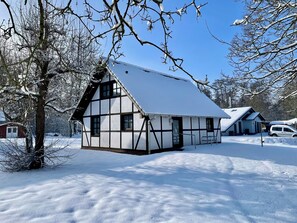 Ein traditionelles Fachwerkhäuschen mit steilem Dach, umgeben von verschneitem Gelände und Bäumen.