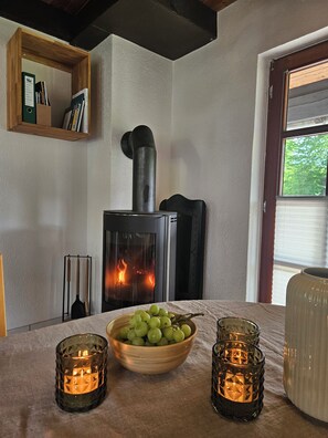 Una habitación con chimenea, un estante de madera con libros, un cuenco de uvas y dos velas encendidas sobre una mesa.