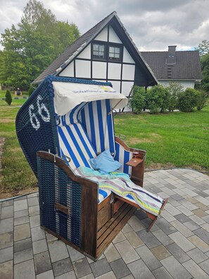 Una silla de playa con un cojín a rayas y un toldo, ubicada en un área pavimentada con una casa al fondo.