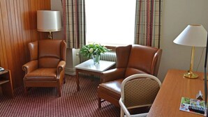 A hotel room with two leather armchairs, a small table with a vase of flowers, a lamp, and a view of the outside through a window with curtains.
