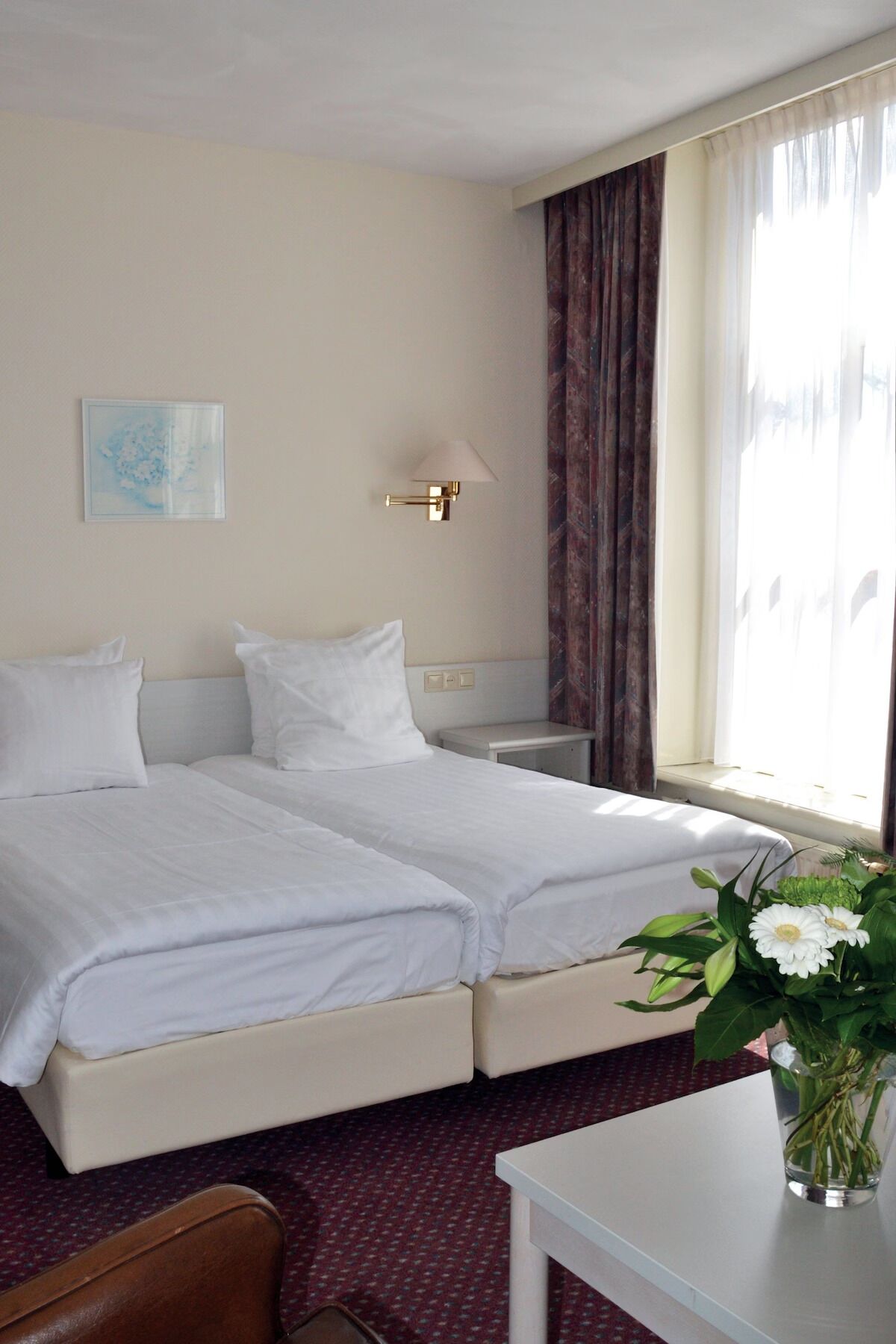 Una habitación de hotel con dos camas, una mesita de noche, una lámpara, un cuadro y un jarrón con flores.