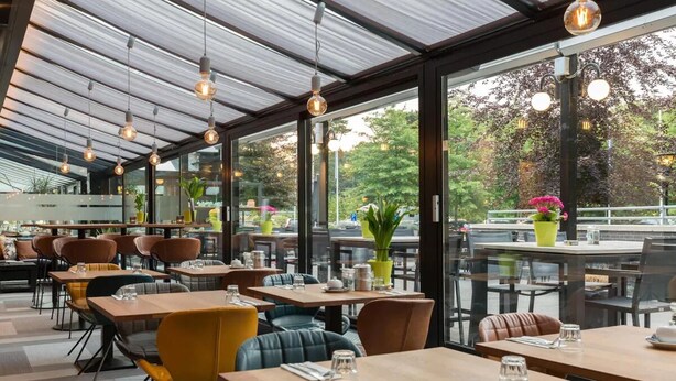 Un restaurante con mesas y sillas, ventanas amplias y lámparas colgantes.