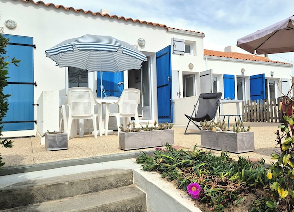 A white building with blue shutters, outdoor seating with chairs and an umbrella, and a garden with plants and a flower.