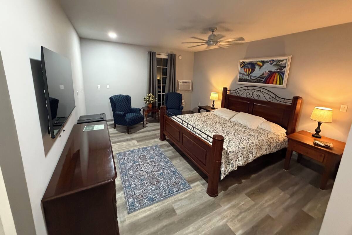 A bedroom with a bed, a desk, a chair, a ceiling fan, a painting, and a window with curtains.