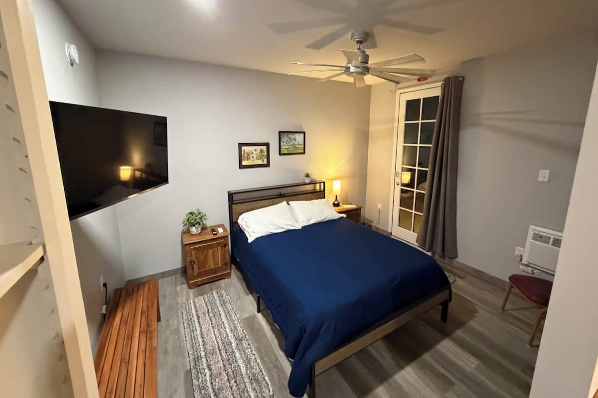 A bedroom with a bed, a ceiling fan, a TV mounted on the wall, a nightstand with a lamp, and a window with curtains.