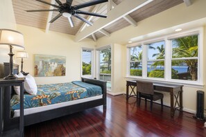 A bedroom with a bed, desk, chair, and large windows with a view of palm trees and the ocean. - Hale Honu 2 Bedroom Home (Kailua-Kona)