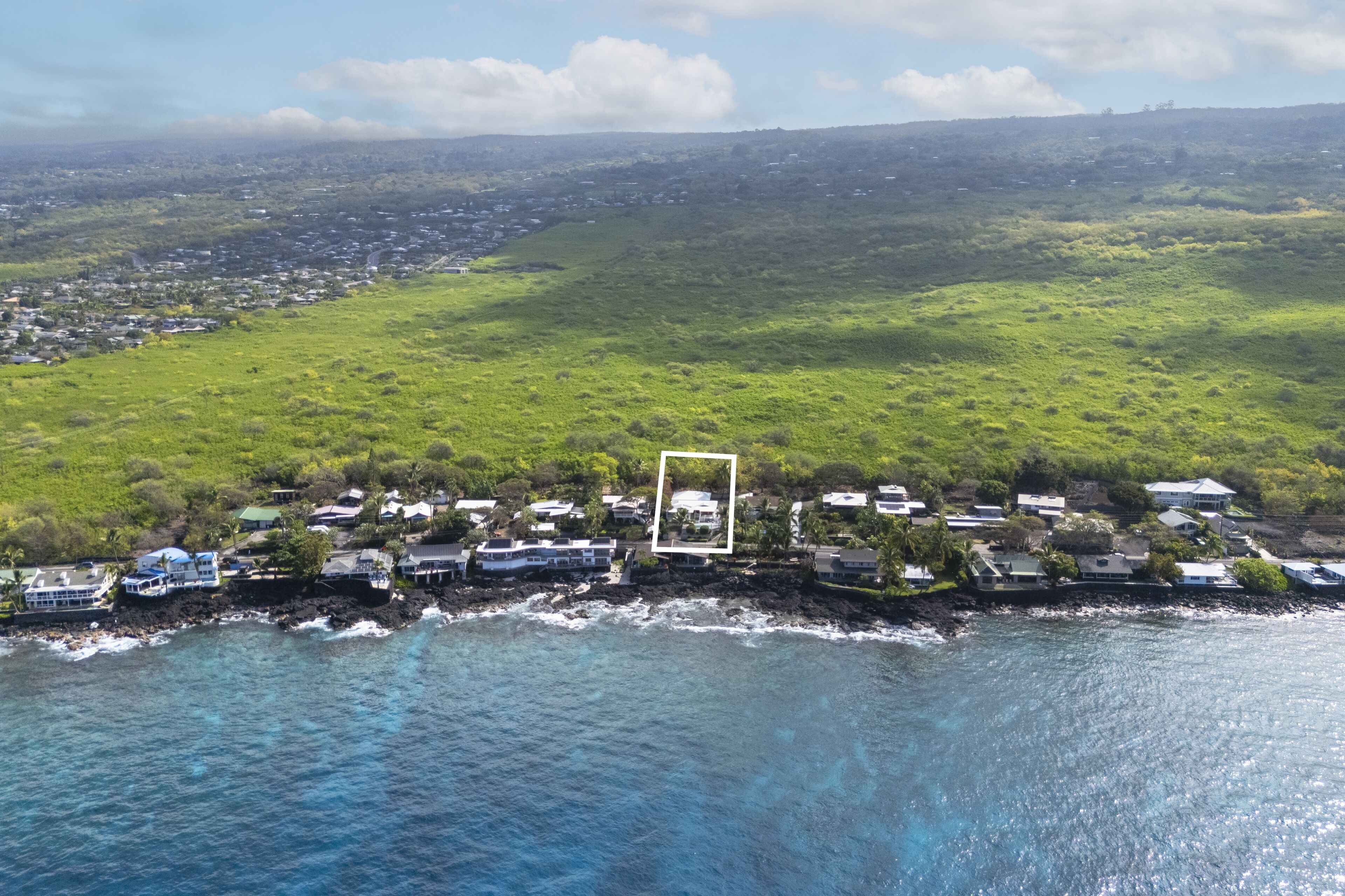 An aerial view of a coastal area with houses along the shoreline, a clear view of the ocean, and a green landscape in the background.