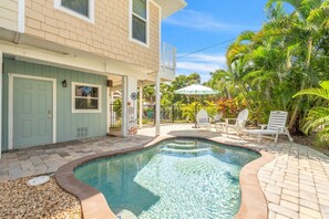 A swimming pool with a tiled deck, a covered patio area with chairs, and a building with a light green door. - Nautical Nest 2 Bedroom Home (Holmes Beach)