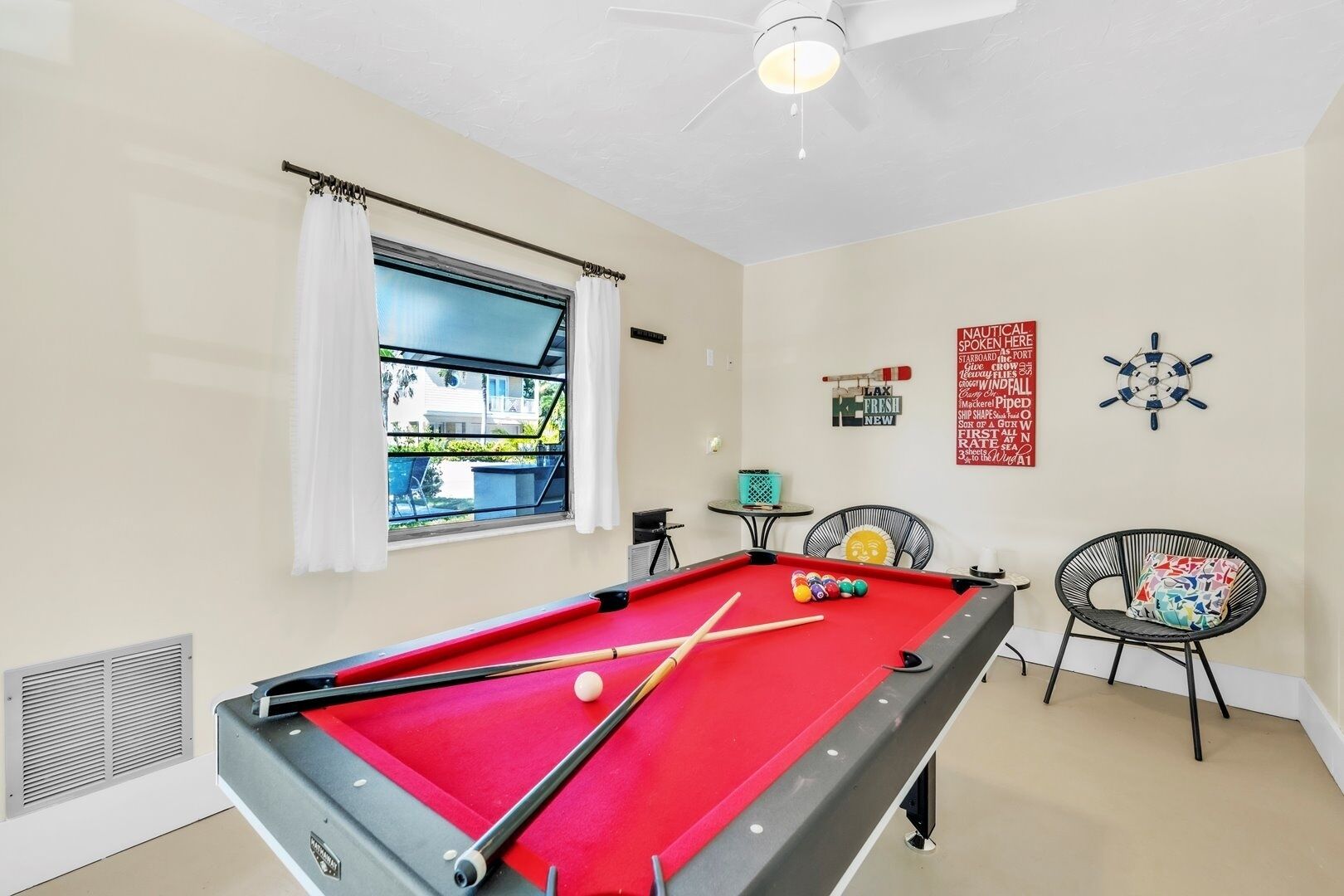 A pool table with balls, a ceiling fan, a window with curtains, a wall clock, and two chairs.
