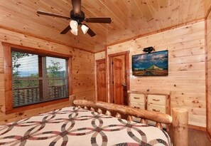 A wooden cabin bedroom with a bed, a ceiling fan, a window with blinds, a door, and a painting on the wall. - Sherwood Love To See You Again 3 Bedroom Cabin (Pigeon Forge)