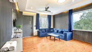 Een moderne zithoek met een blauwe bank, een houten salontafel en een groot raam met uitzicht op bomen.