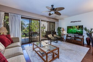 A living room with a glass coffee table, a sofa, a television, and a ceiling fan. - Maui Kamaole B102 1 Bedroom Condo (Kihei)