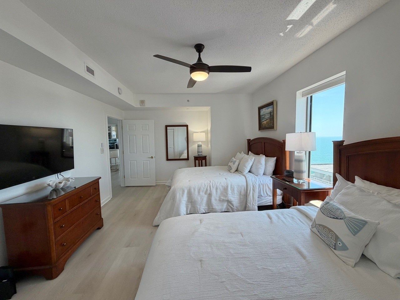 A hotel room with two beds, a television, a ceiling fan, and a view of the sea.