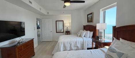 A hotel room with two beds, a television, a ceiling fan, and a view of the sea.