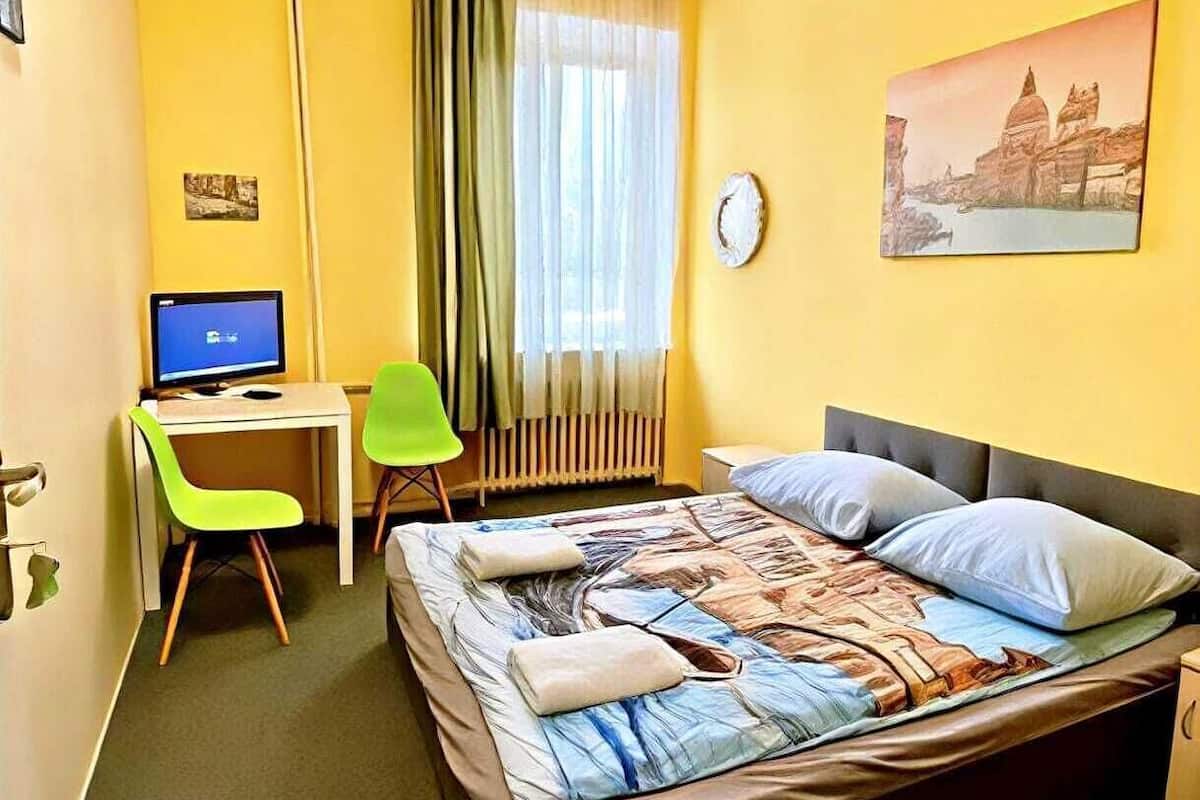 Une chambre d’hôtel comprenant un lit, un bureau avec un ordinateur, une chaise, une fenêtre avec des rideaux et une horloge murale.