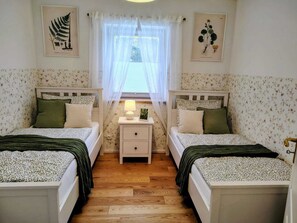 Une chambre avec deux lits, une table de chevet, une fenêtre avec des rideaux légers et des œuvres d’art murales sur le thème de la botanique.