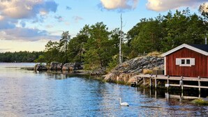 A wooden dock extending into calm water, a red cabin with a white window, surrounded by trees and rocks. - Idyllic house by the sea with motorboat - Scandinavian, child-friendly, modern (Loftahammar)