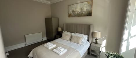 A hotel room with a bed, a radiator, a wardrobe, a nightstand with a lamp, and a potted plant.