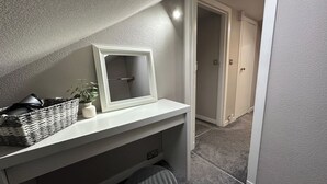 Ein Badezimmer mit weißem Waschtisch, einem Spiegel, einem Korbwäscheschrank, einer Topfpflanze und einem gestreiften Hocker.
