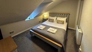 Ein Schlafzimmer mit schrägen Wänden, einem Bett mit gesteppter Matratze, zwei Kissen, einem Nachttisch und einem Beistelltisch mit Lampe, sowie einem Fenster.