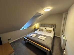 Un dormitorio con techo inclinado, una cama con colchón acolchado, dos almohadas, una mesita de noche y una lámpara en un mueble auxiliar, y una ventana.