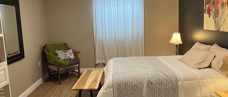 A bedroom with a bed, a chair, a small table, a lamp, and a painting on the wall.