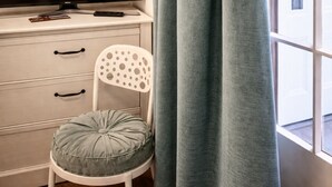 Habitació amb televisió sobre una tauleta blanca, una cadira blanca amb coixí i una finestra amb cortines.