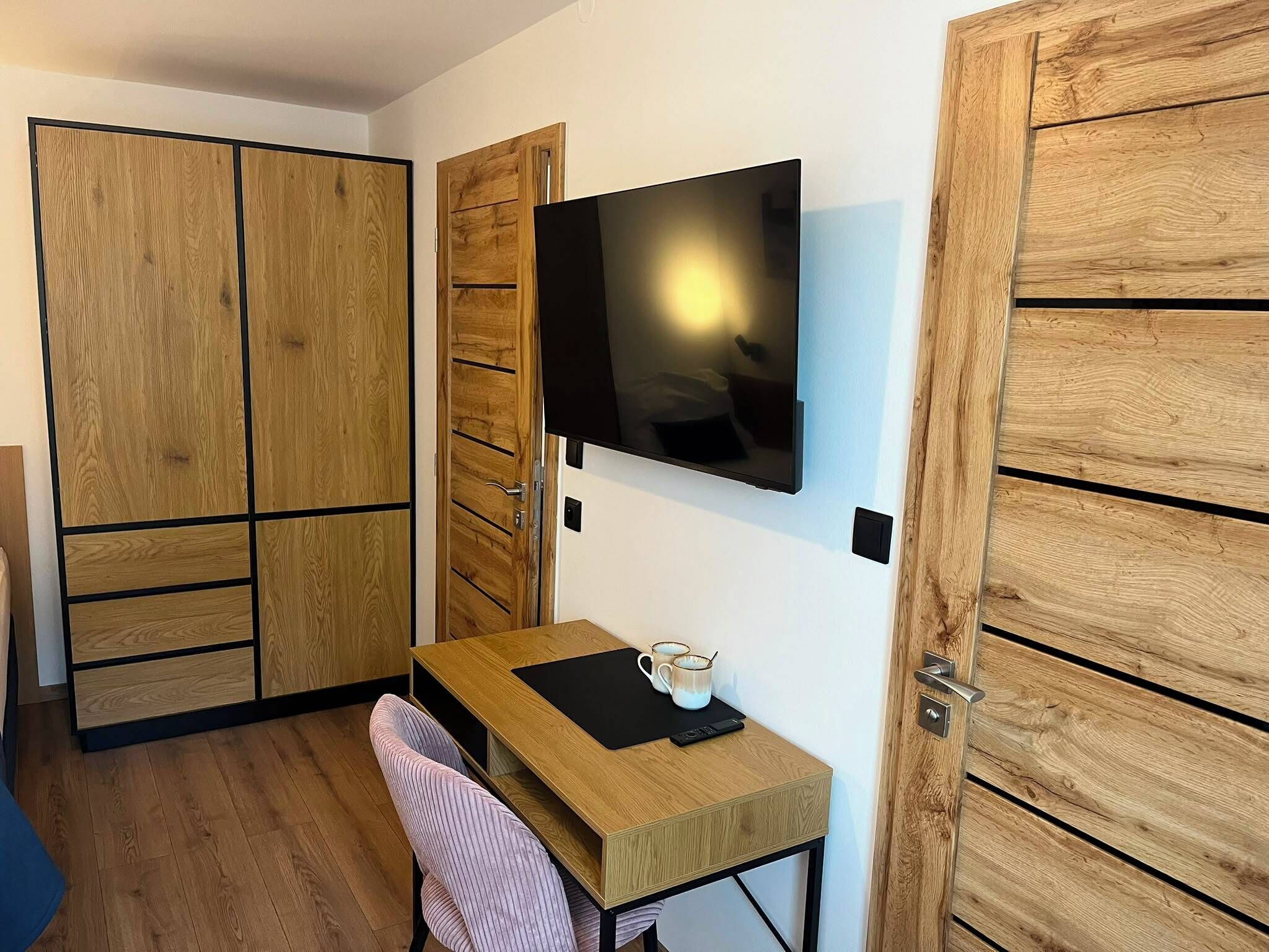 A small room with a wooden desk, a chair, a television mounted on the wall, and wooden wardrobes.