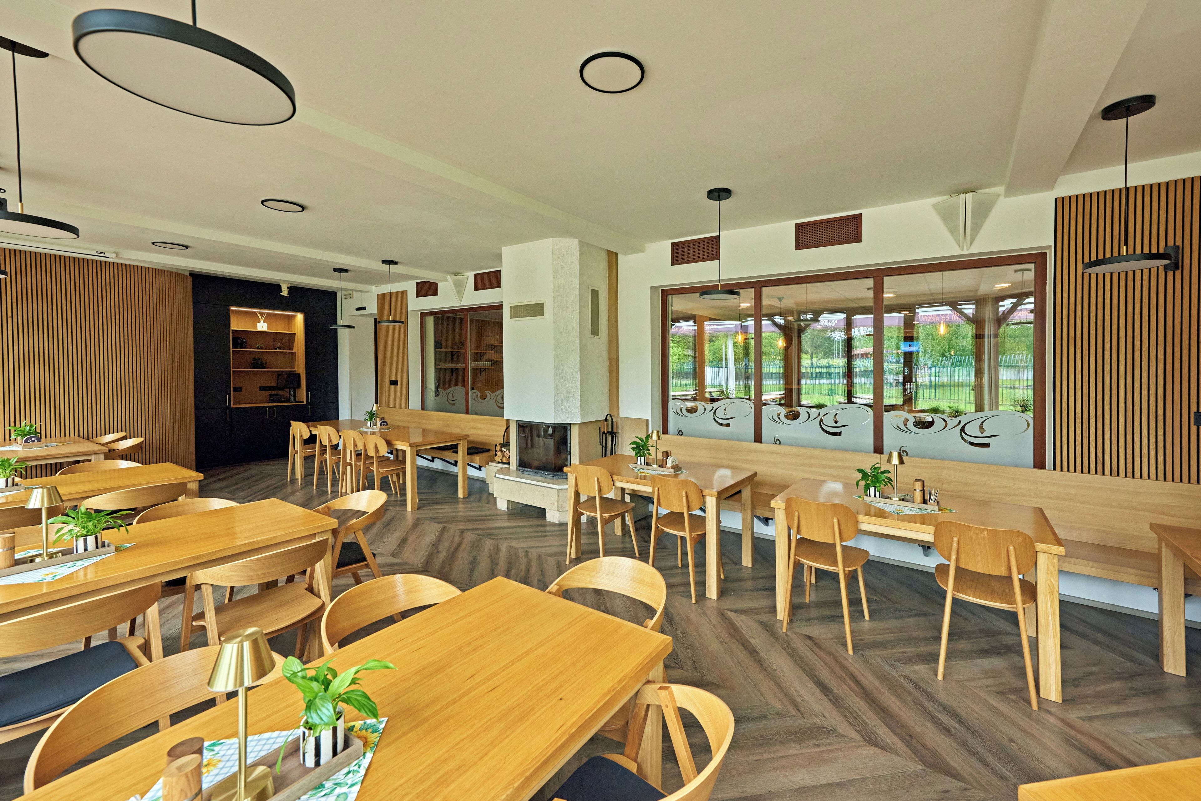 A modern dining area with wooden tables and chairs, a fireplace, and large windows offering a view of the outdoors.
