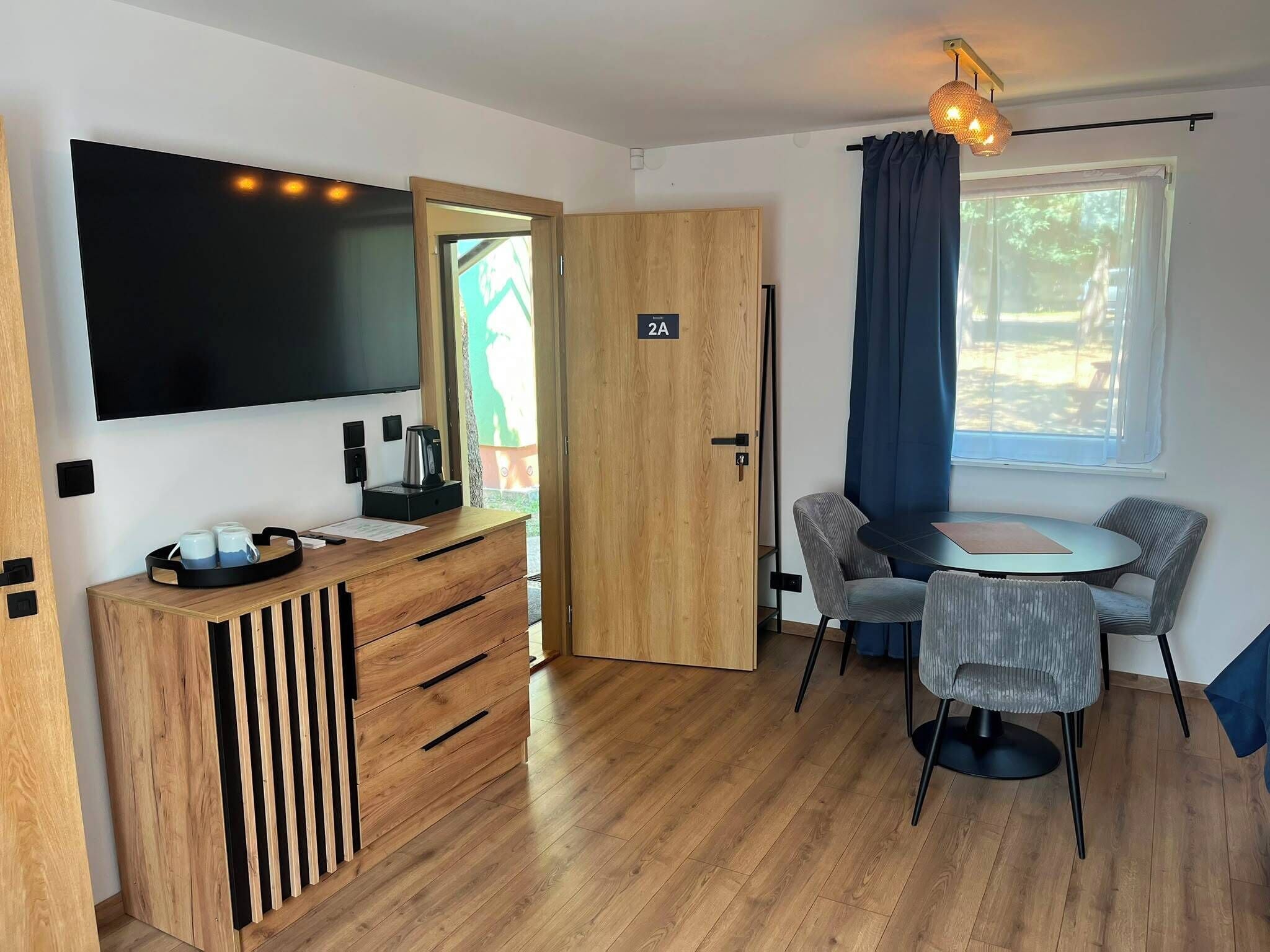 A modern room with a flat-screen TV, a wooden desk with a kettle, a dining area with a table and chairs, and a large window with curtains.