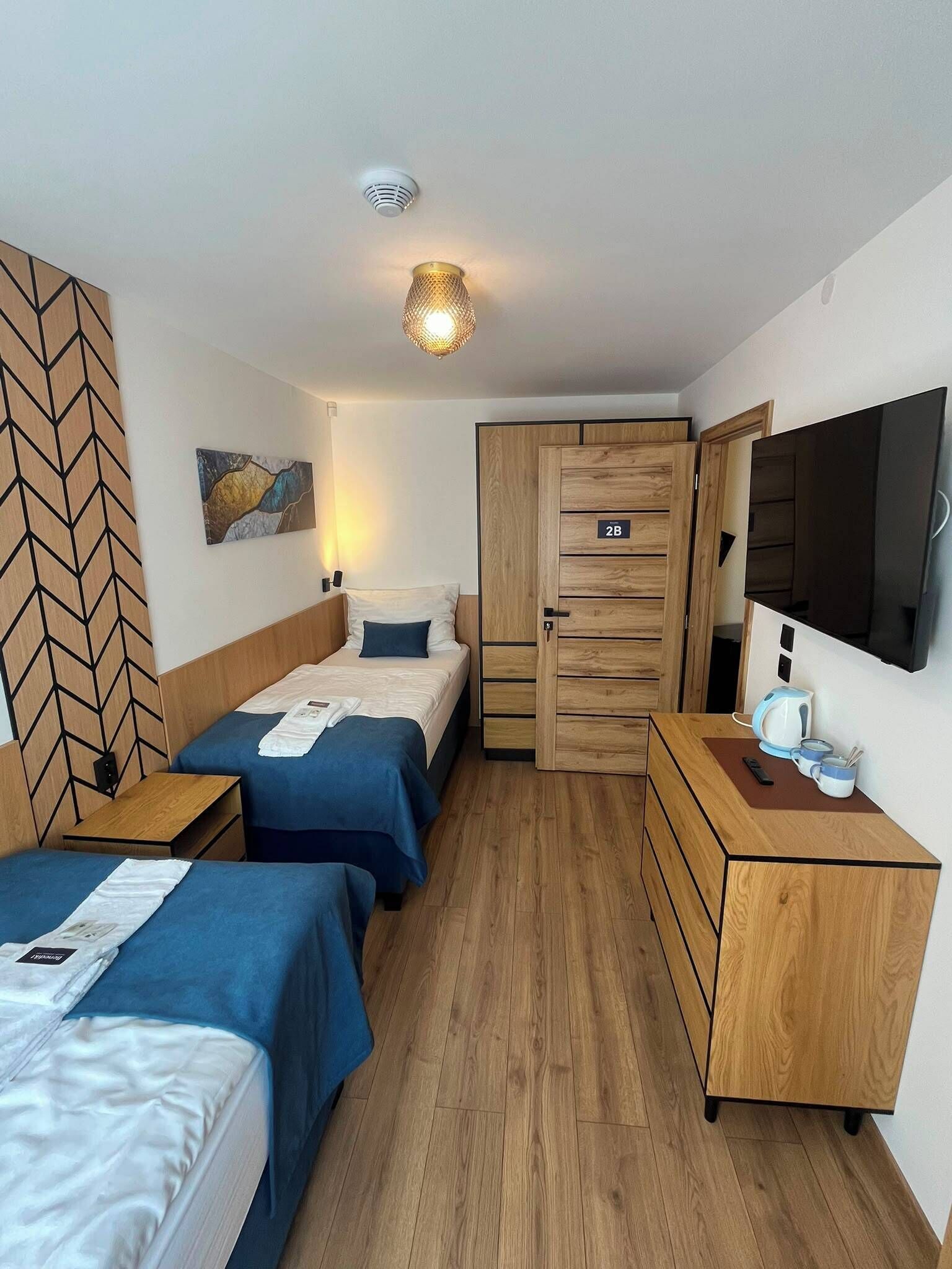 Ein Hotelzimmer mit zwei Betten, einem Holzschrank, einem Schreibtisch mit Wasserkocher und einem Flachbildfernseher.