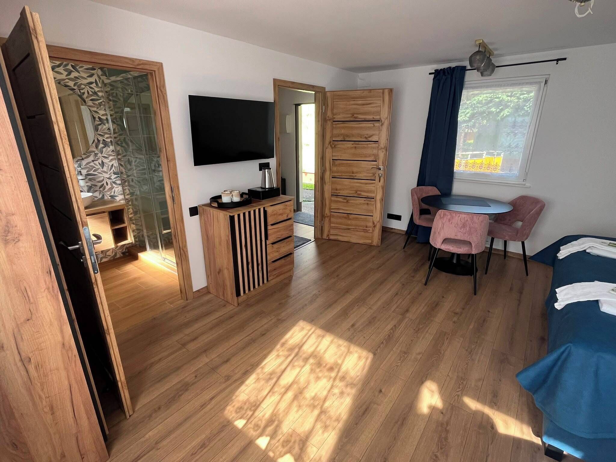 A room with a wooden floor, a TV mounted on the wall, a small table with chairs, and a bed with a blue cover.