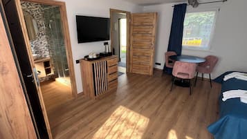 A room with a wooden floor, a TV mounted on the wall, a small table with chairs, and a bed with a blue cover.