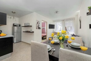 Et moderne køkken med køleskab, mikroovn og et spiseområde med et bord dækket til to personer, pyntet med en vase med gule blomster og glas med appelsinjuice.