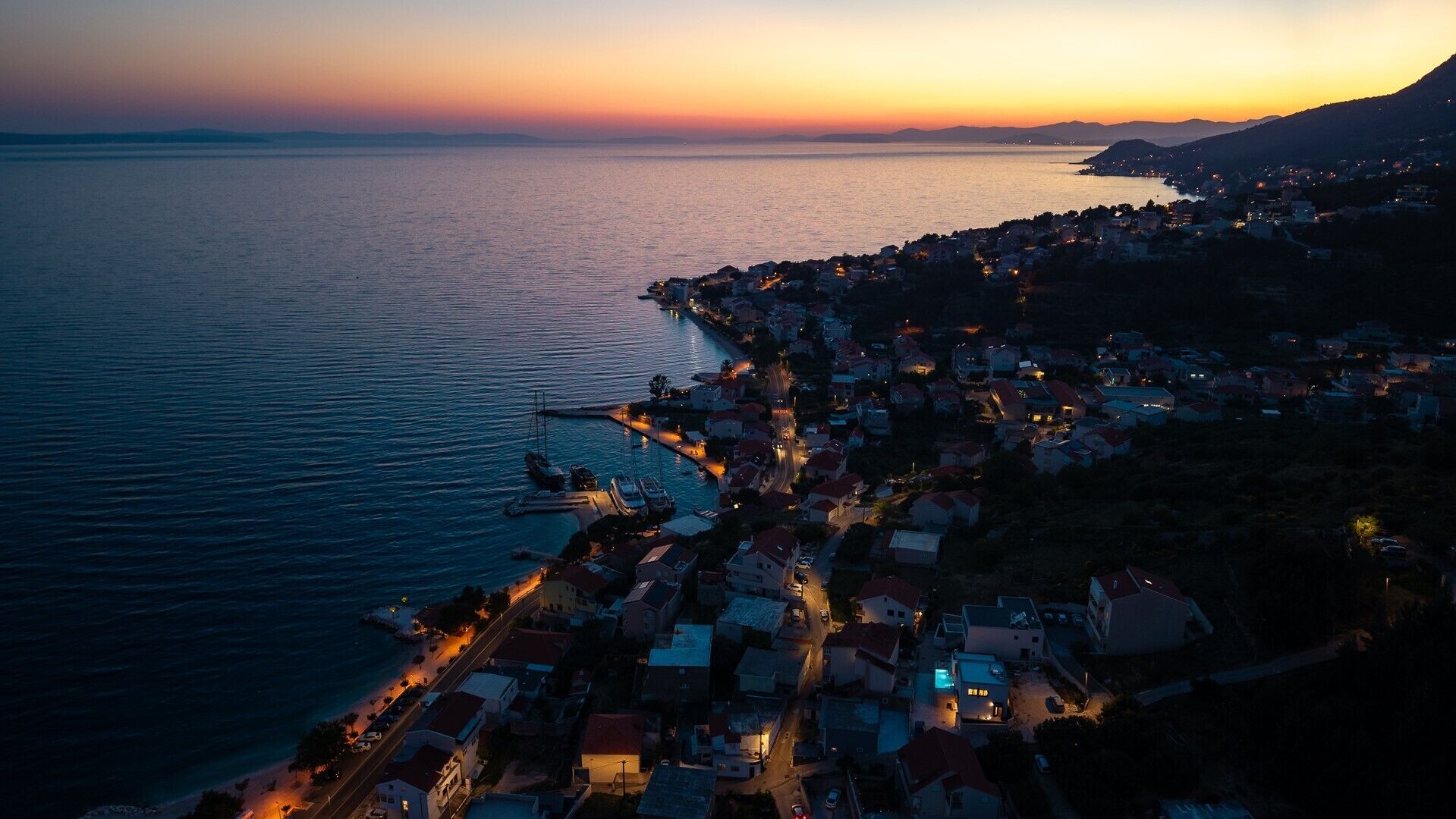Vista aérea de uma cidade costeira ao entardecer, com um porto de iates, edifícios residenciais e um relevo montanhoso ao fundo.
