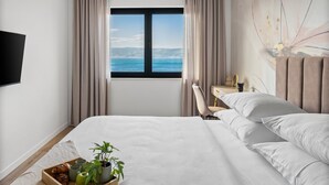 Hotelkamer met een bed, bureau, stoel, raam met uitzicht op zee en een dienblad met eten en een plant.